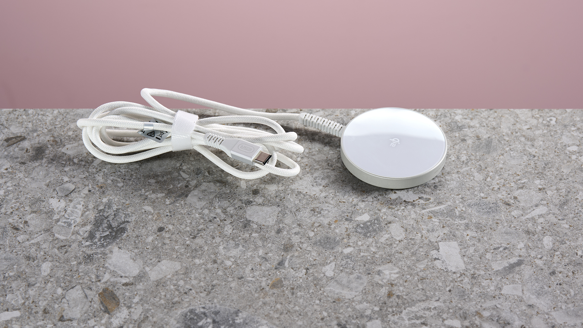 ESR Qi2 Mini Wireless Charger sits on a light grey stone-effect surface against a pink background. The folded and secured charging cable extends from the left of the charging pad.