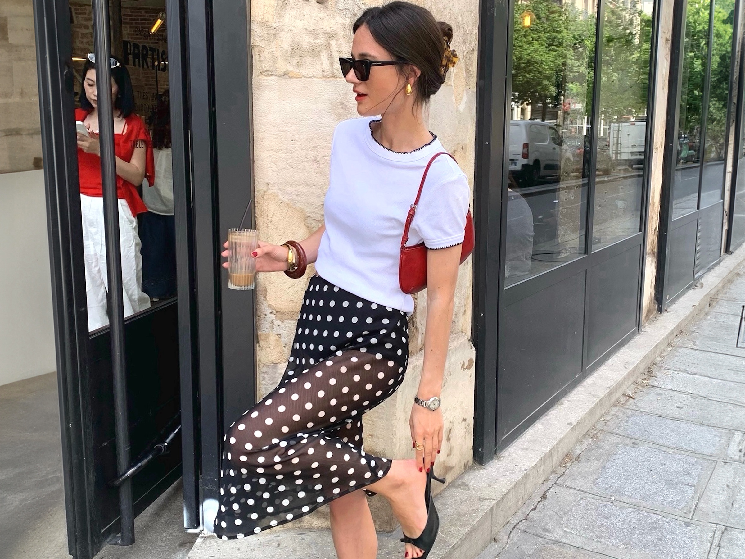 The French-style influencer of Parisian poses with rib glass outside a cafe in Paris, wearing claw clips on one side, black sunglasses, thick gold earrings, white contrast hem T-shirt, mini red shoulder bag, bracelet, polka dots, polka dots skirt, simple silver watch and black kitten moore sandals,