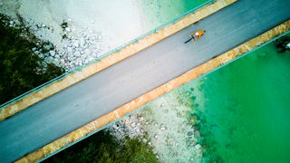An aerial view of a person cycling on a bridge over turquoise water, surrounded by rocky shore and lush greenery