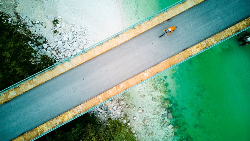 An aerial view of a person cycling on a bridge over turquoise water, surrounded by rocky shore and lush greenery