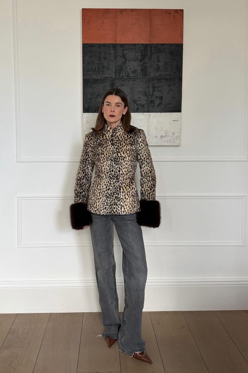 A woman standing inside a home in front of a white wall with modern art wearing a dalmatian print pony hair jacket with gray straight-leg jeans and brown pointed pumps.