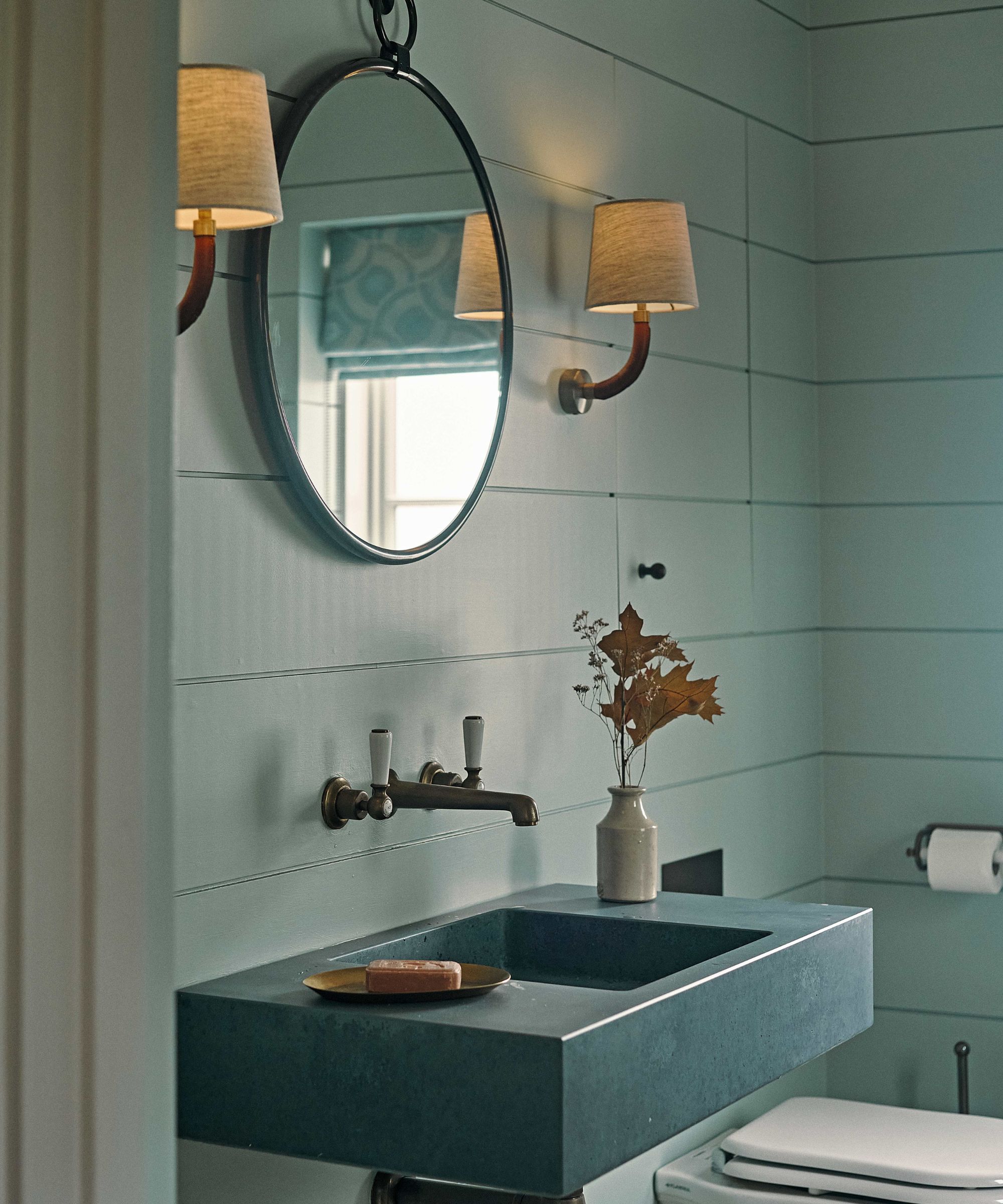 Blue panelled bathroom with blue stone sink, circular mirror and wall lights with cream shades