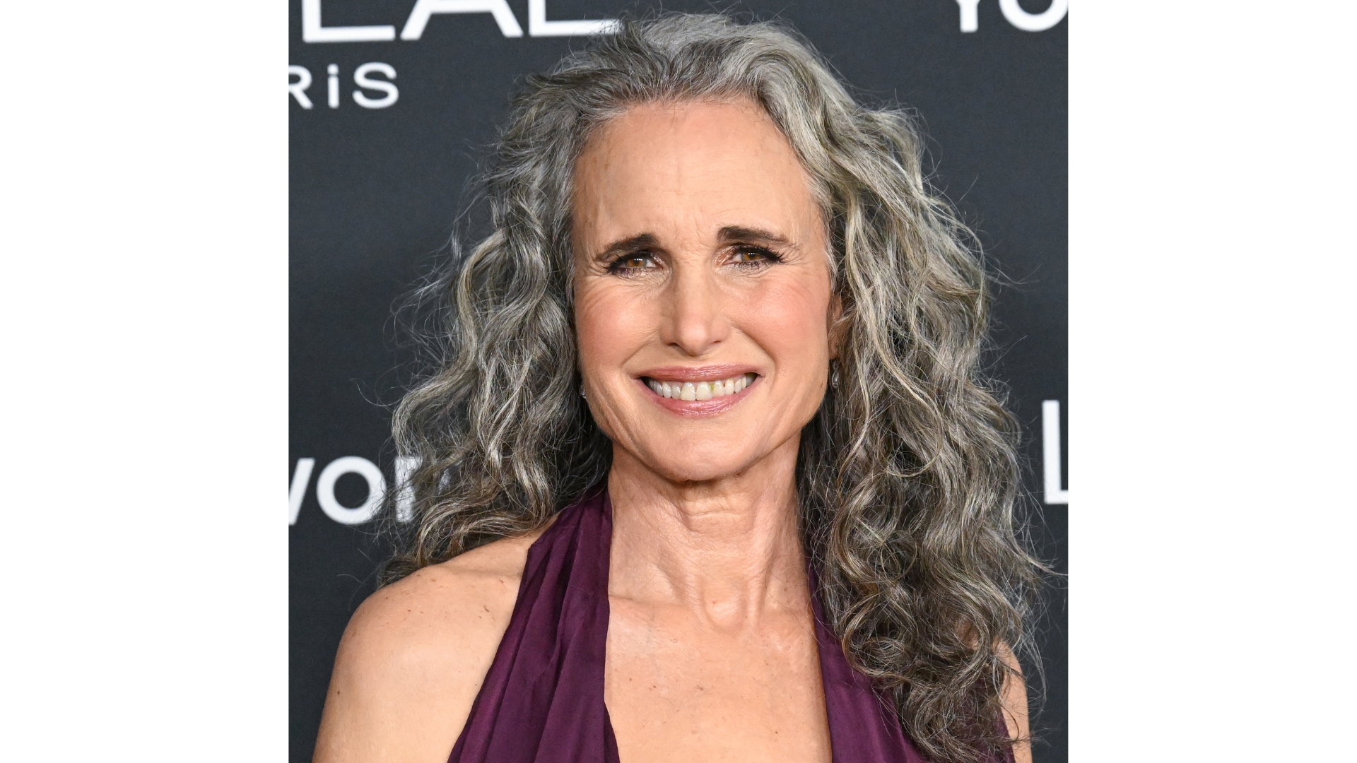 Image of Andie MacDowell smiling at the camera with curly long grey hair and wearing a purple dress