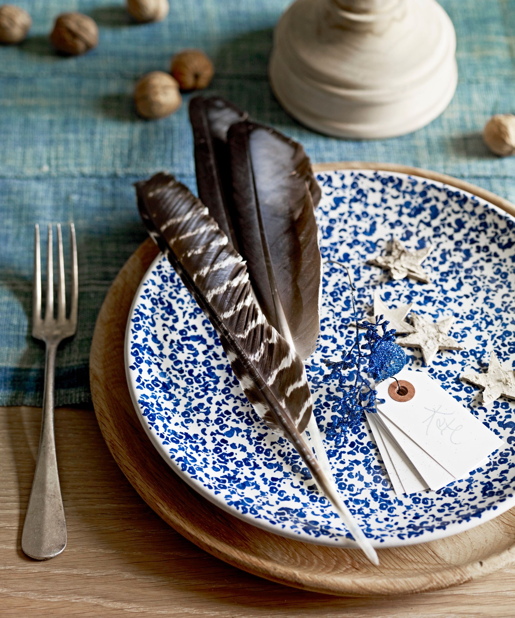 Christmas place setting with feathers, stars and nuts on a blue and white speckled plate and wooden charger