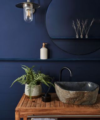 Dark bathroom with indigo wall tiles and wall, with round mirror and organic stone sink