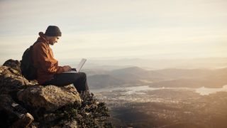 A young hiker using his laptop while sitting on top of a mountain.