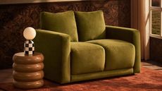 A compact green sofa on a burnt orange rug next to a sculptural brown side table with a checkered table lamp on it