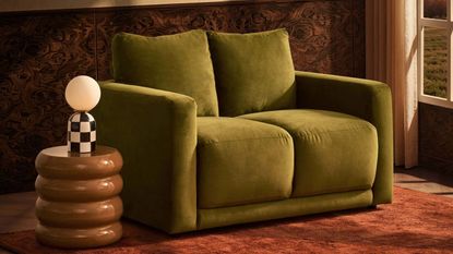A compact green sofa on a burnt orange rug next to a sculptural brown side table with a checkered table lamp on it