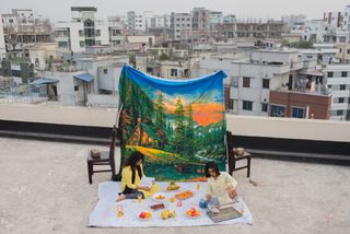 Two people sit on a rooftop picnic setup surrounded by a colorful feast, with a vibrant nature-themed backdrop