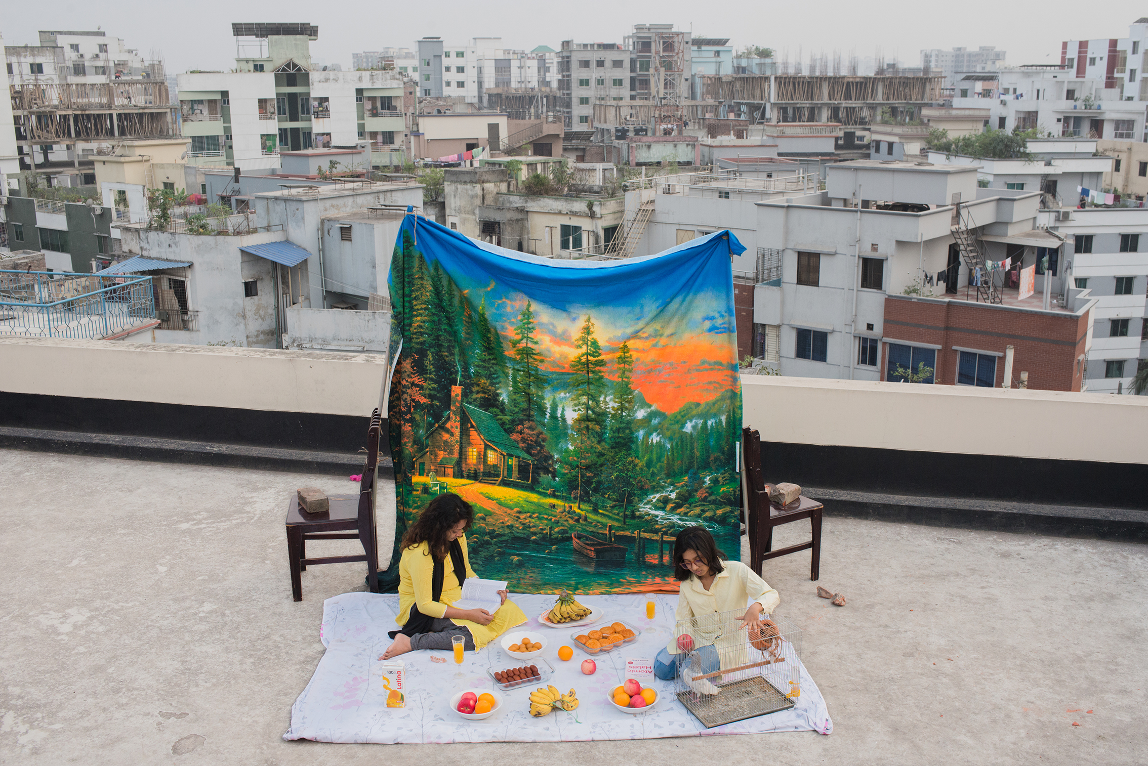 Two people sit on a rooftop picnic setup surrounded by a colorful feast, with a vibrant nature-themed backdrop
