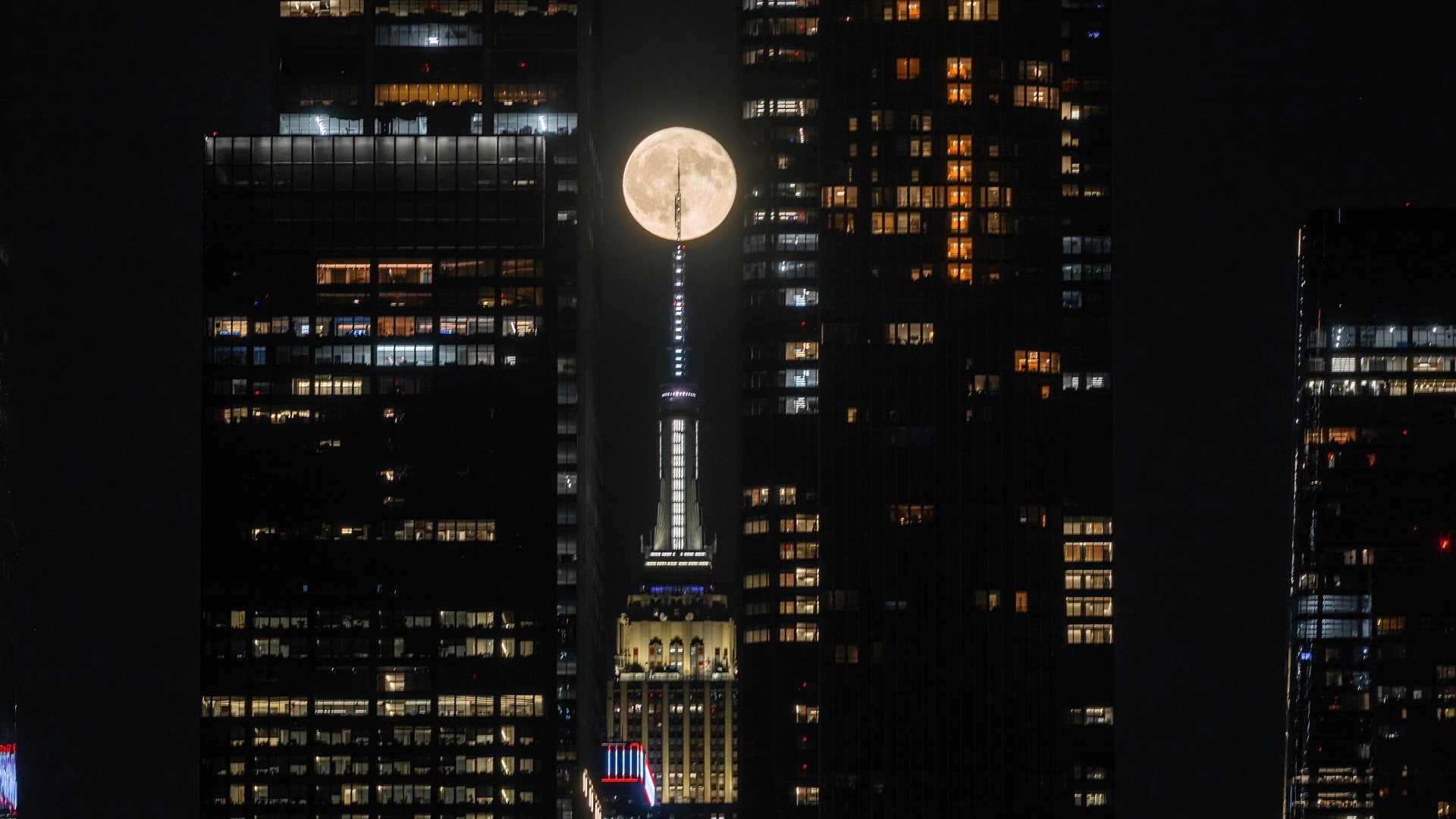 A full moon is pictured against a night sky between two skyscrapers. The tip of a third skyscraper — the iconic Empire State Building — can be seen directly below the lunar disk, with its point appearing to spear the moon.