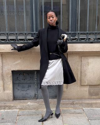 Sylvie mus wears a black blazer, black sweater, belt, white lace skirt, gray tights, and black slingbacks.