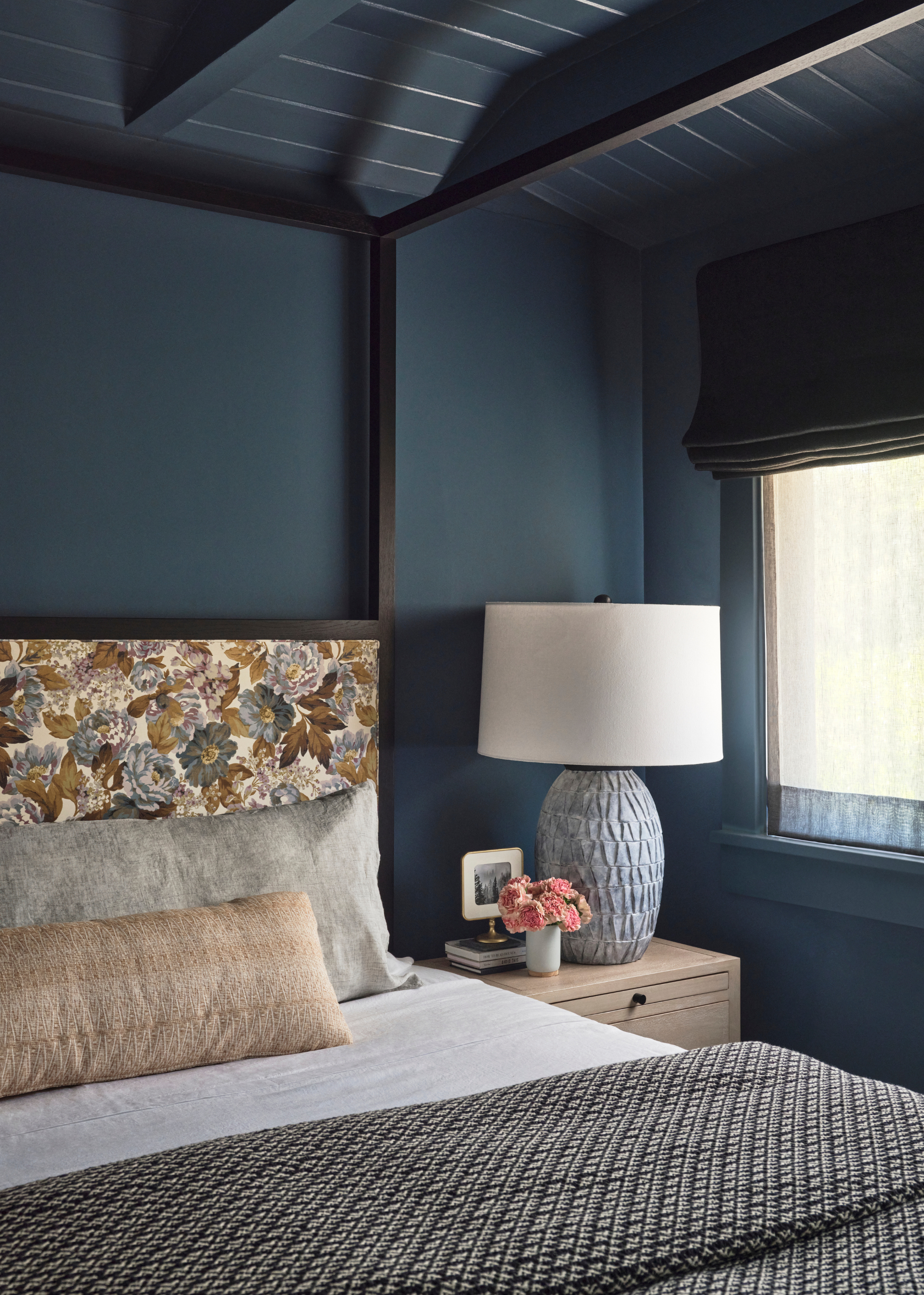 A blue bedroom with a patterned headboard, a tan bolster pillow, a wood side table with a lamp and vase, and a patterned duvet
