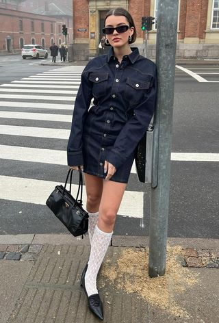 A woman's jean dress outfit with button-front minidress styled with lace knee-high socks, black pointed pumps, and a black Prada bag.