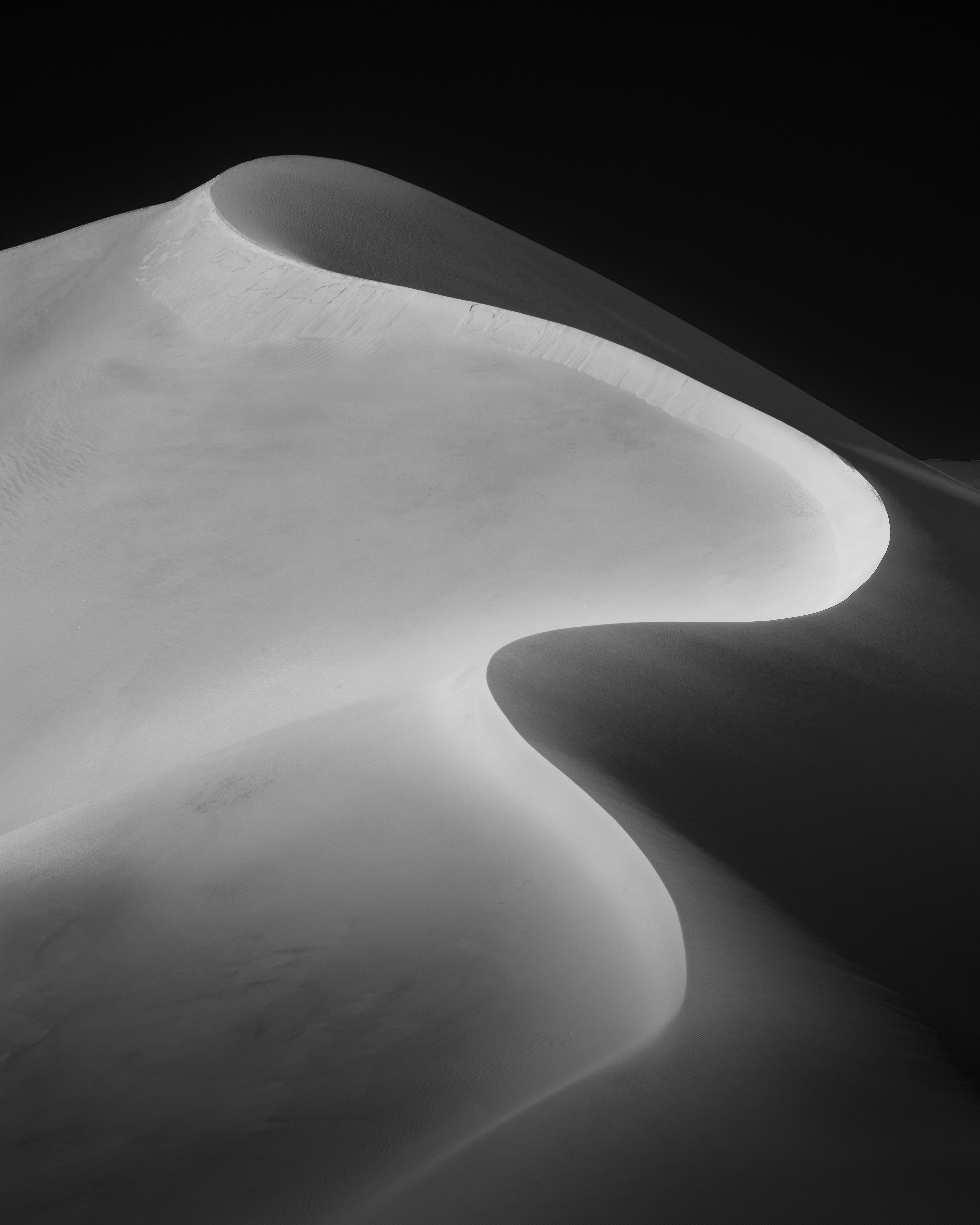 A striking black-and-white photograph captures the smooth, curving ridge of a massive sand dune against a dark background, emphasizing light and shadow
