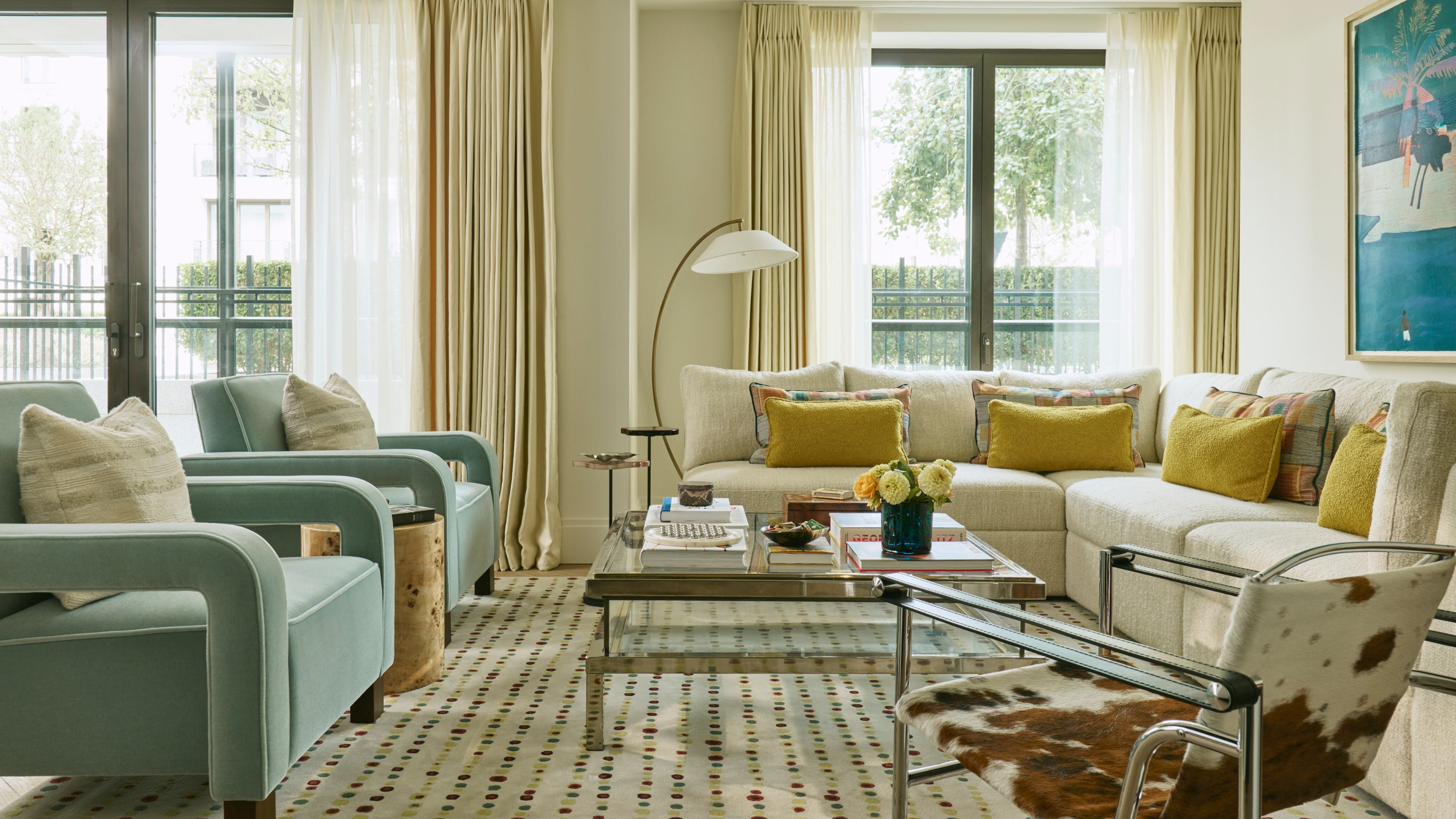a cream londonopen-plan apartment living area with a large cream sectional, large windows, blue armchairs and pops of yellow with a cow print chair