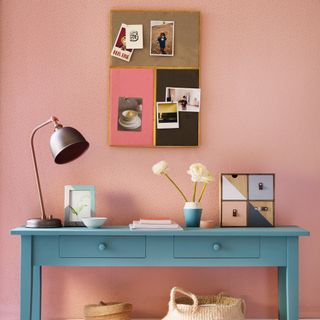 White and pink wall and rug, with corkboard and baby blue sideboard covered in objects in shades of blue, pink, yellow, and orange
