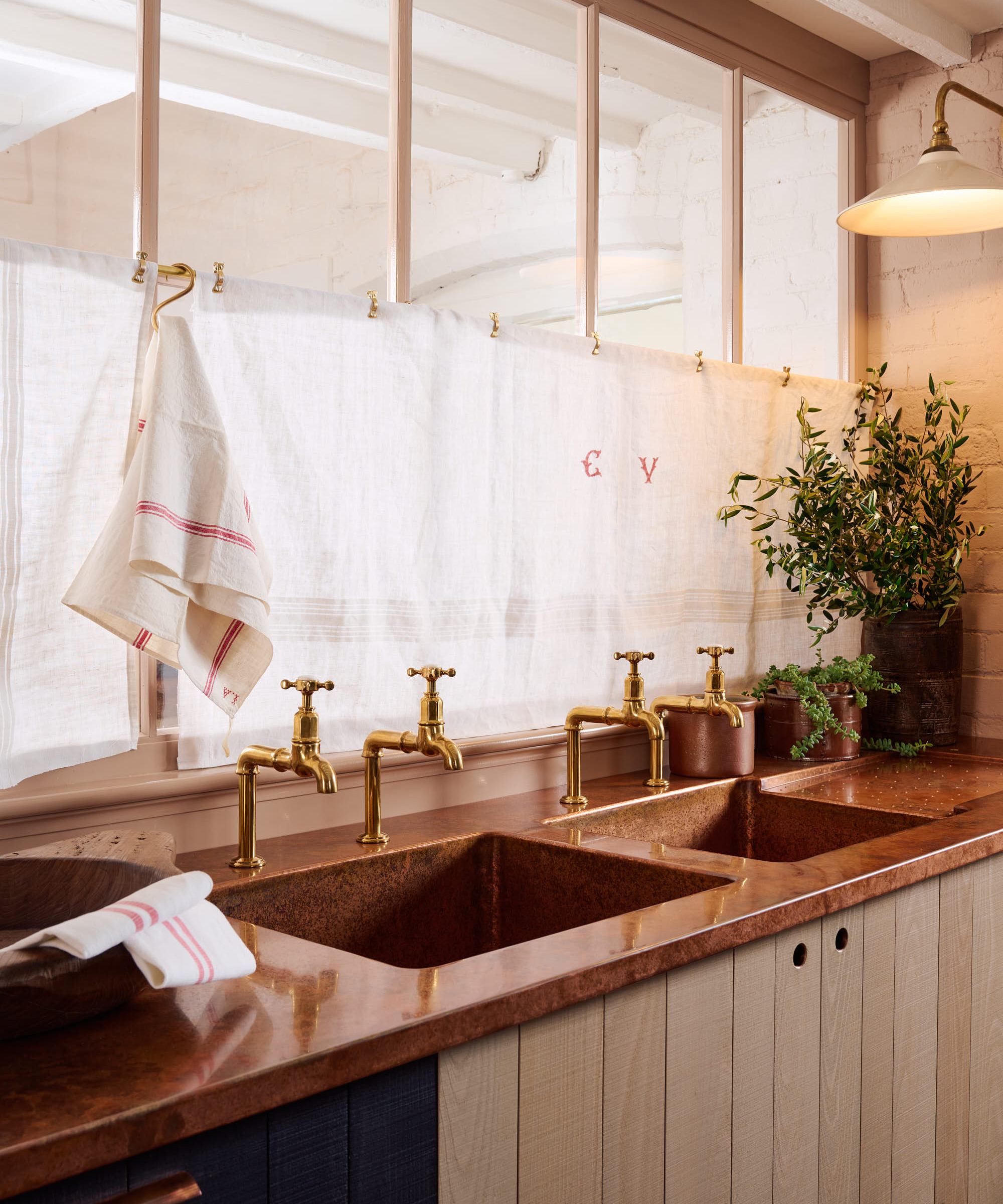 internal window in kitchen covered with cafe style curtains above double sink area inset into copper worktop with wooden cabinet doors below