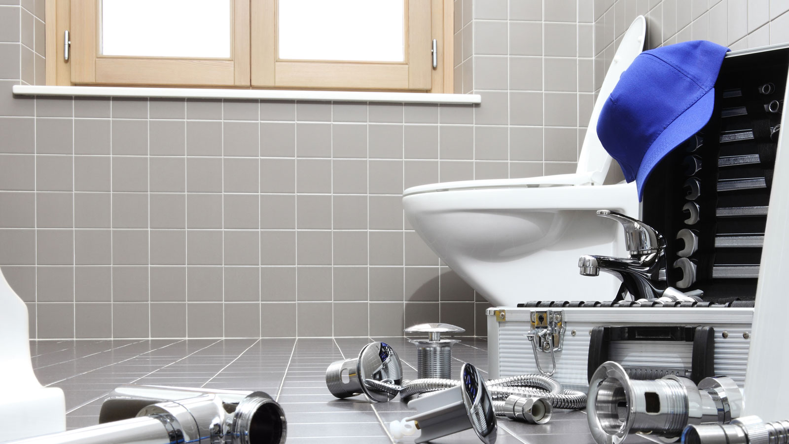 Plumbing parts on floor in front of white toilet in bathroom with square grey wall and floor tiles