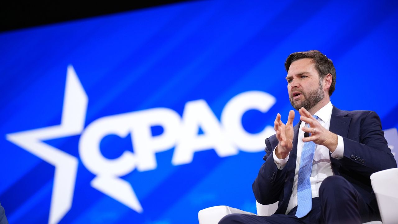 JD Vance, seated, at the CPAC convention. He is wearing a dark-blue suit with a white buttondown shirt and a light-blue tie. He is gesturing with his hands