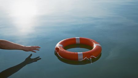 A hand reaches for an orange life ring floating on the surface of calm water.