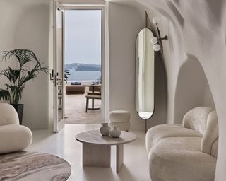 A white room with a velvet couch, oval mirror, large plant and open door looking out onto the sea