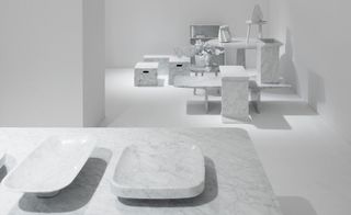 In the foreground: white marble style dishes of differing shapes. In the backdrop is various types of furniture including side tables, benches, boxes and vases.