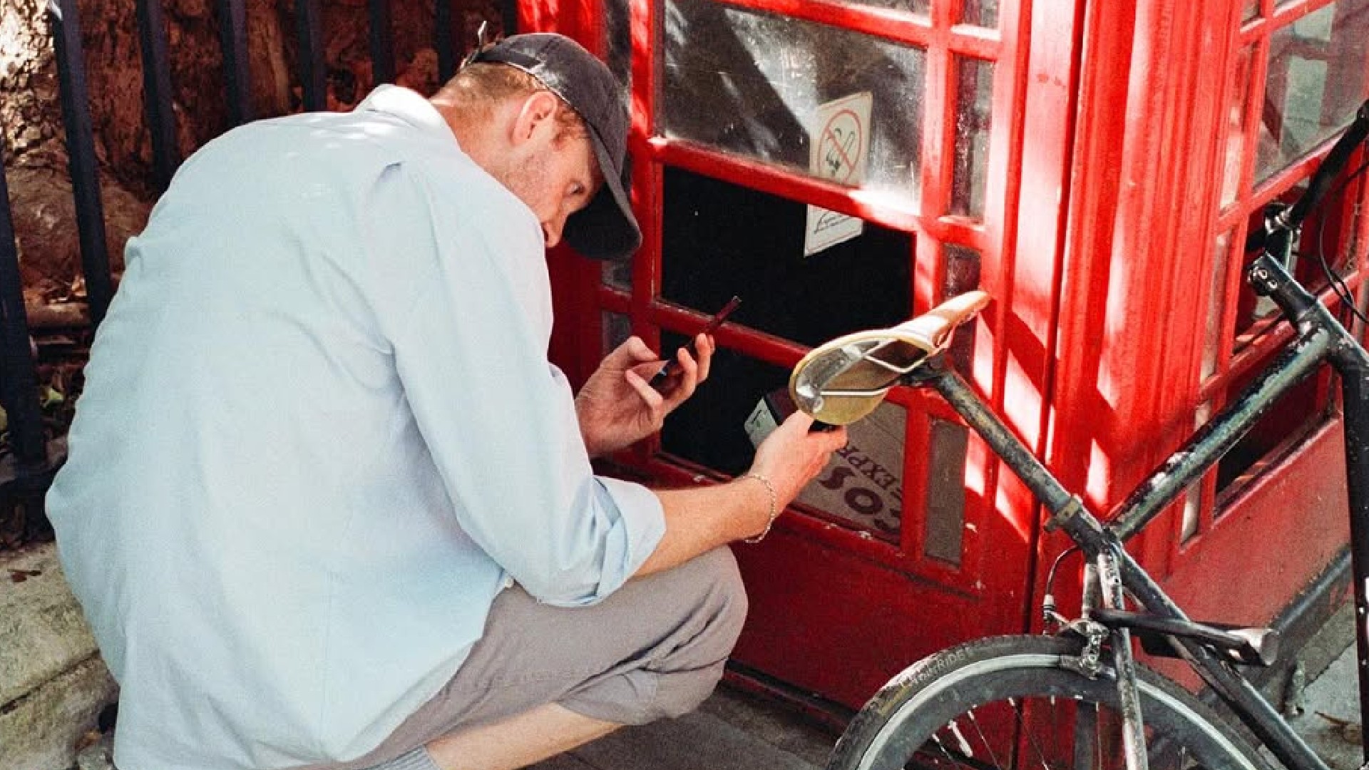 A man wearing a light blue shirt and a baseball cap is crouching down next to a red telephone booth, appearing to be examining something inside or on the ground while holding a small black object and a bottle.
