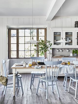 A calming white kitchen idea with pale blue painted chairs, gray wooden flooring and white cabinetry and pendant lighting.