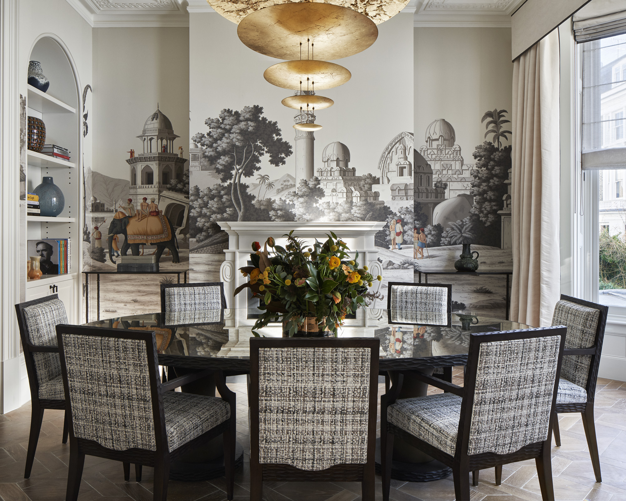 A dining table decor idea with round dark wood table, brass chandelier in a modern style, grey mural wallpaper and a floral centerpiece