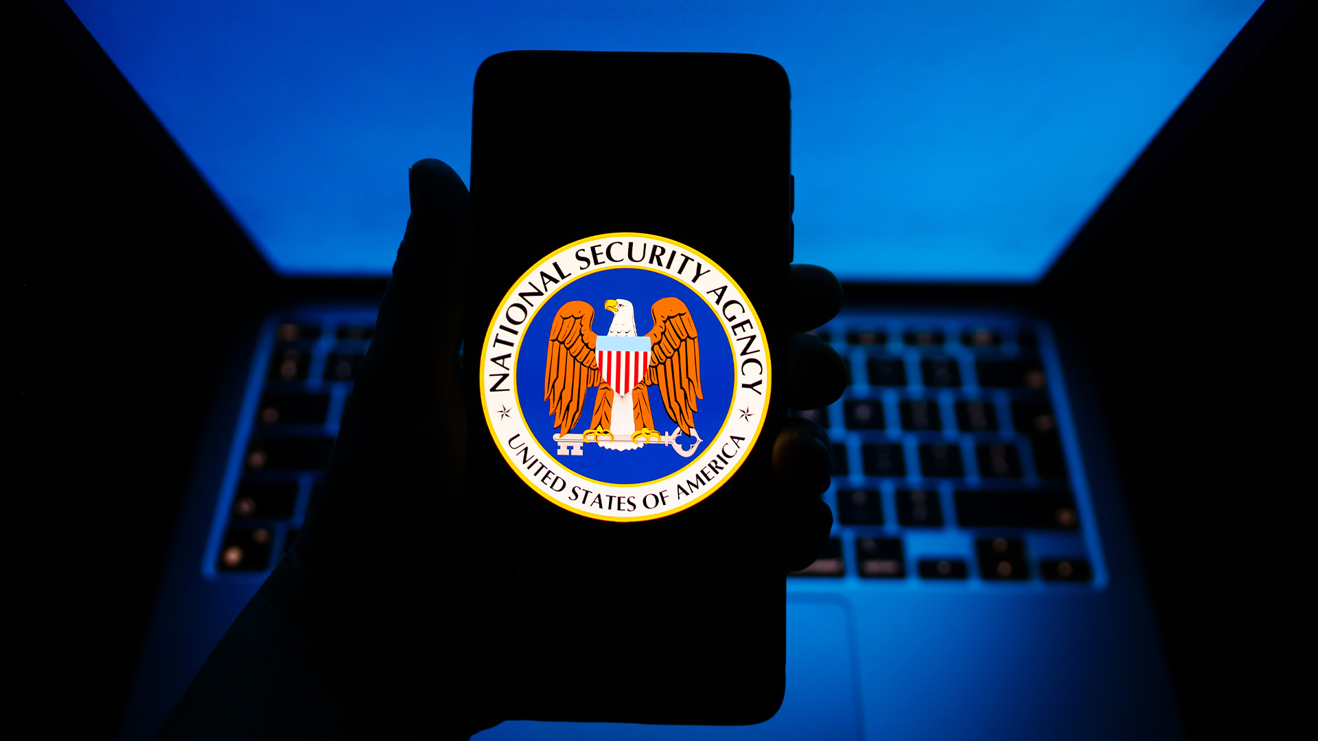 Logo and insignia of the United States National Security Agency (NSA) pictured on a smartphone screen held in hand above a laptop keyboard and screen.