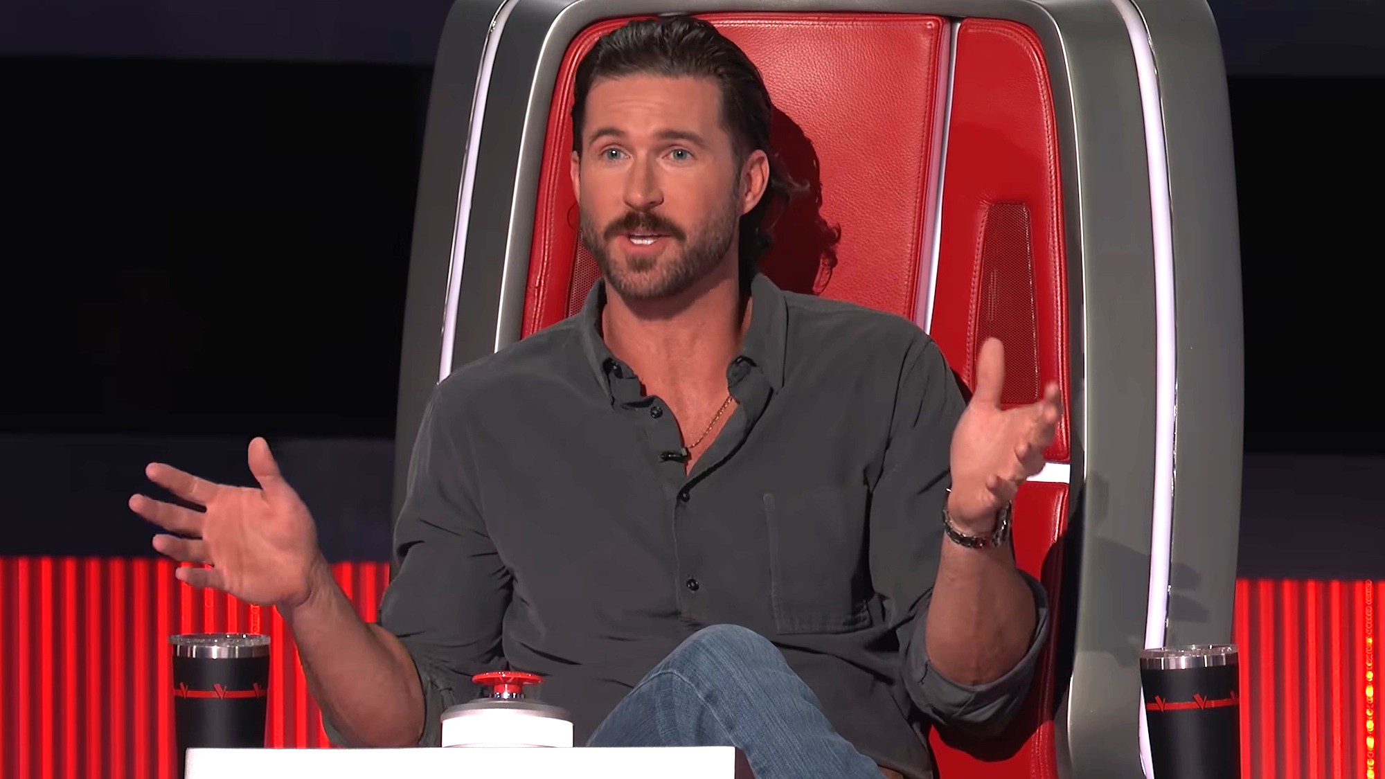 Riley Green is shown sitting in his Big Red Chair on The Voice Season 30, wearing a gray button-down shirt and jeans as he talks to an artist on stage.
