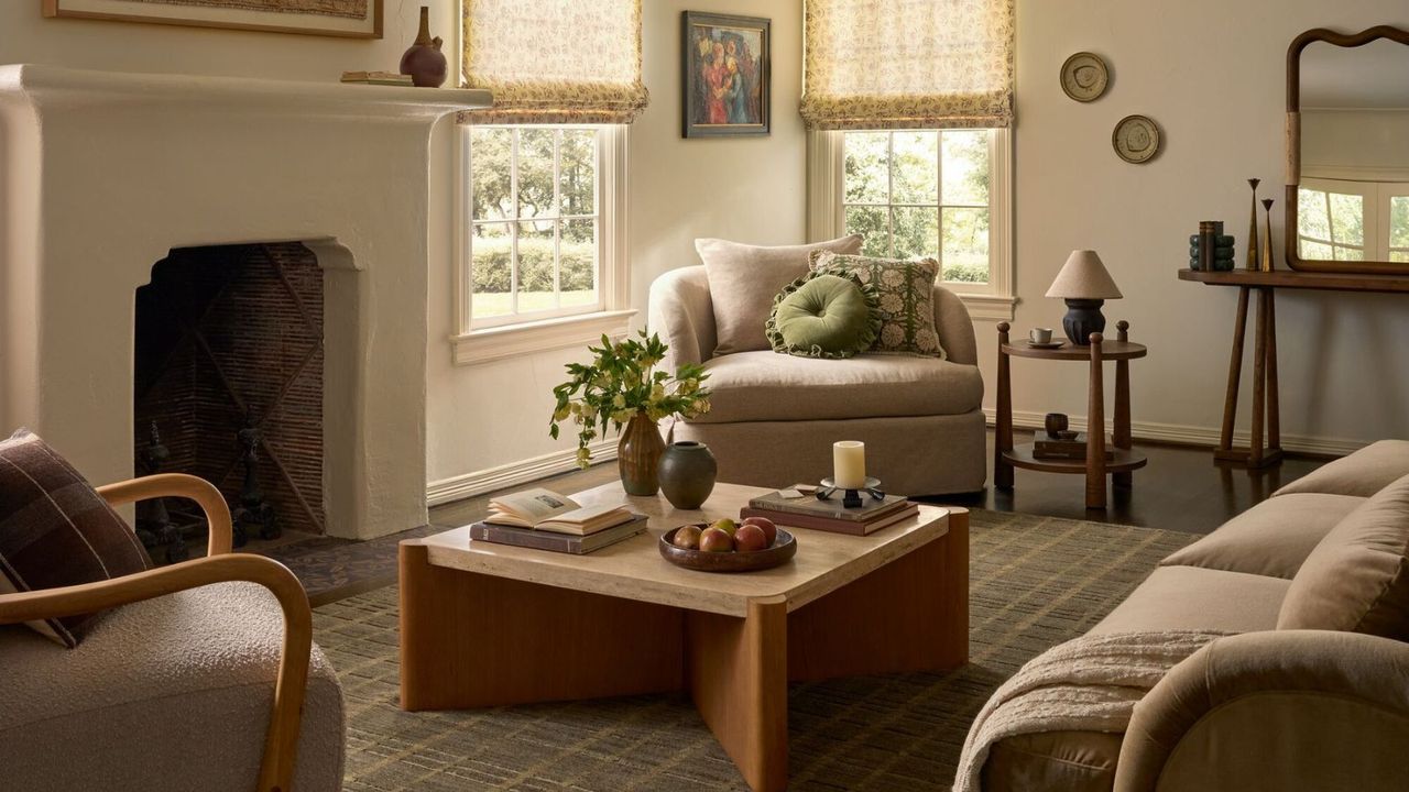 A cozy but bright cream living room with cream upholstered chairs, cuddle seats and mid-centrury wooden coffee table, side table and sideboard. There is a stone mantel and sheer window treatments in view, as well as a checkered area rug