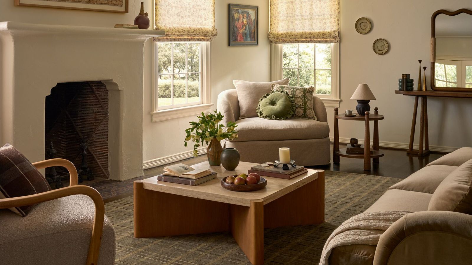 A cozy but bright cream living room with cream upholstered chairs, cuddle seats and mid-centrury wooden coffee table, side table and sideboard. There is a stone mantel and sheer window treatments in view, as well as a checkered area rug