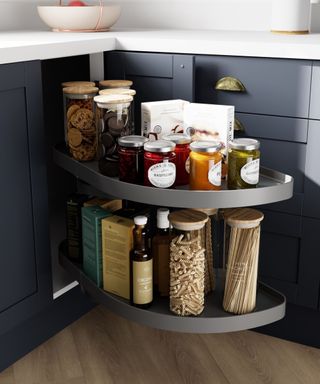 A dark blue corner kitchen cabinet with two-tiered pull-out storage is neatly organised with glass jars filled with pasta, sauces, cookies, and spices. The cabinet has brass cup handles, and the wooden floor adds warmth to the space.