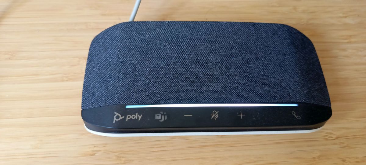 A grey, black and white Poly Sync 10 speakerphone sitting on a brown wooden desk