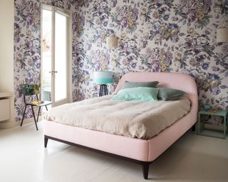 A pink double bed on white floorboards in front of purple floral wallpaper