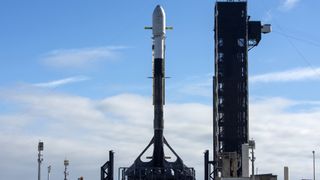 A black and white SpaceX rocket with the Intuitive Machines IM-2 moon lander on the launch pad under a blue sky with clouds.