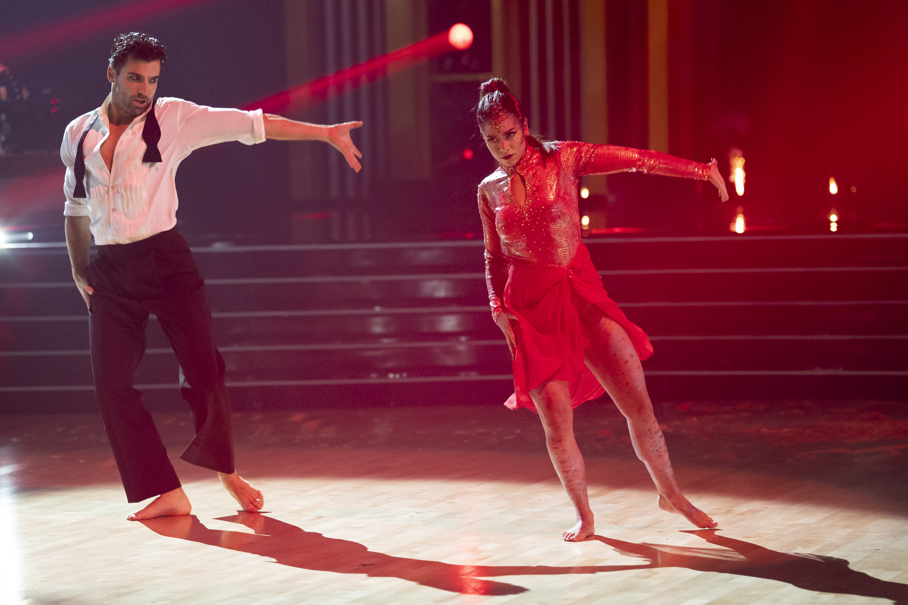JAN RAVNIK and JENNIFER AFFLECK performing a dance wearing a suit shirt and black pants and a red dress under red lighting on season 34 of dancing with the stars