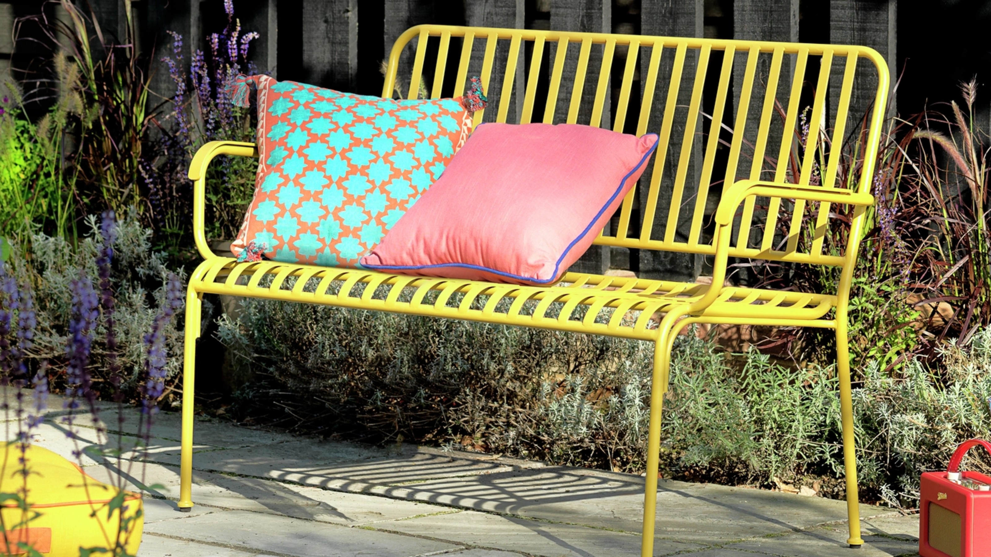 Habitat Indu 2 Seater Metal Garden Bench in Yellow with two colourful garden cushions in a sunny garden