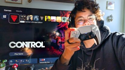 A photo of a person holding a DualSense controller in front of a TV displaying the PS5 homescreen