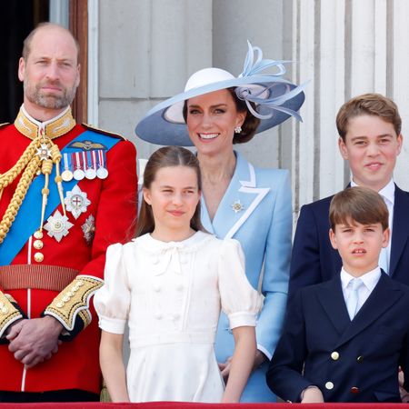 Prince William wears red military uniform and has a bald head and a beard, and stands next to wife Princess Middleton who wears a baby blue coat dress and matching hat, and their three kids