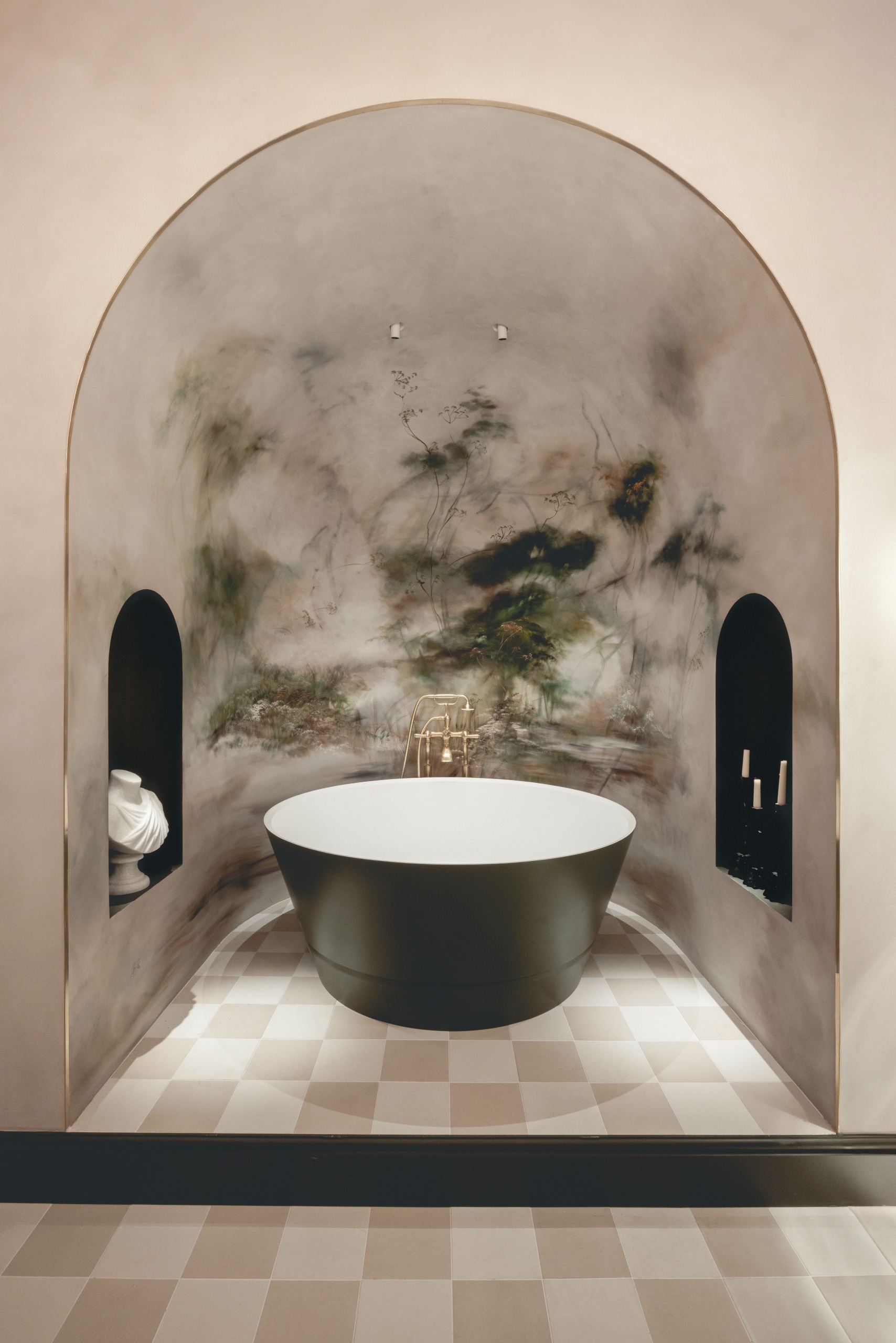 A detail of a bathroom decorated with checkered beige tiles, an abstract, nature-inspired dome with two alcoves, and a soaking bathtub in dark green with brass fixtures.