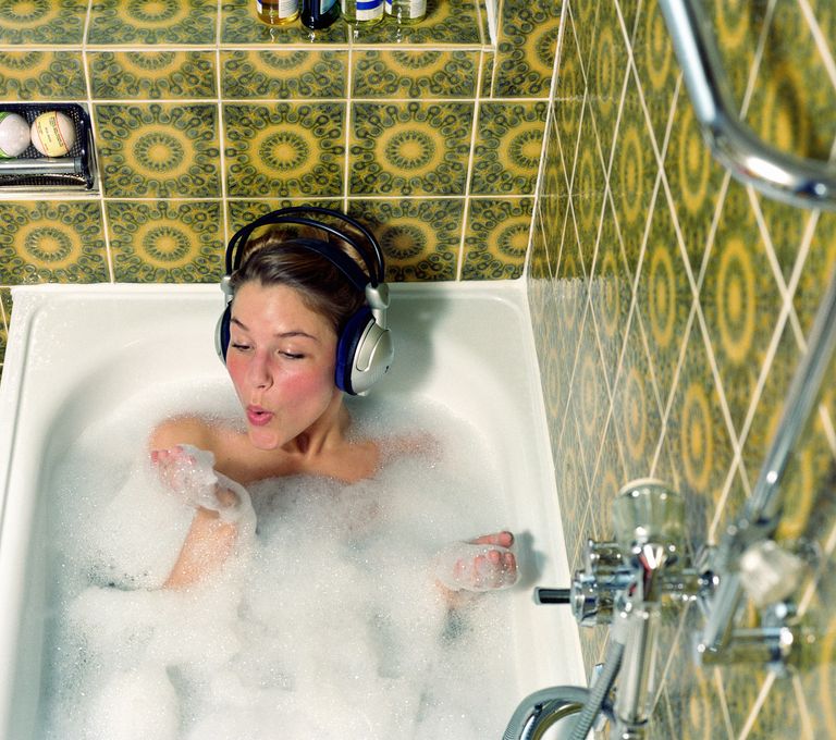 Young woman wearing headphones in bath blowing foam from hand, self-care day ideas