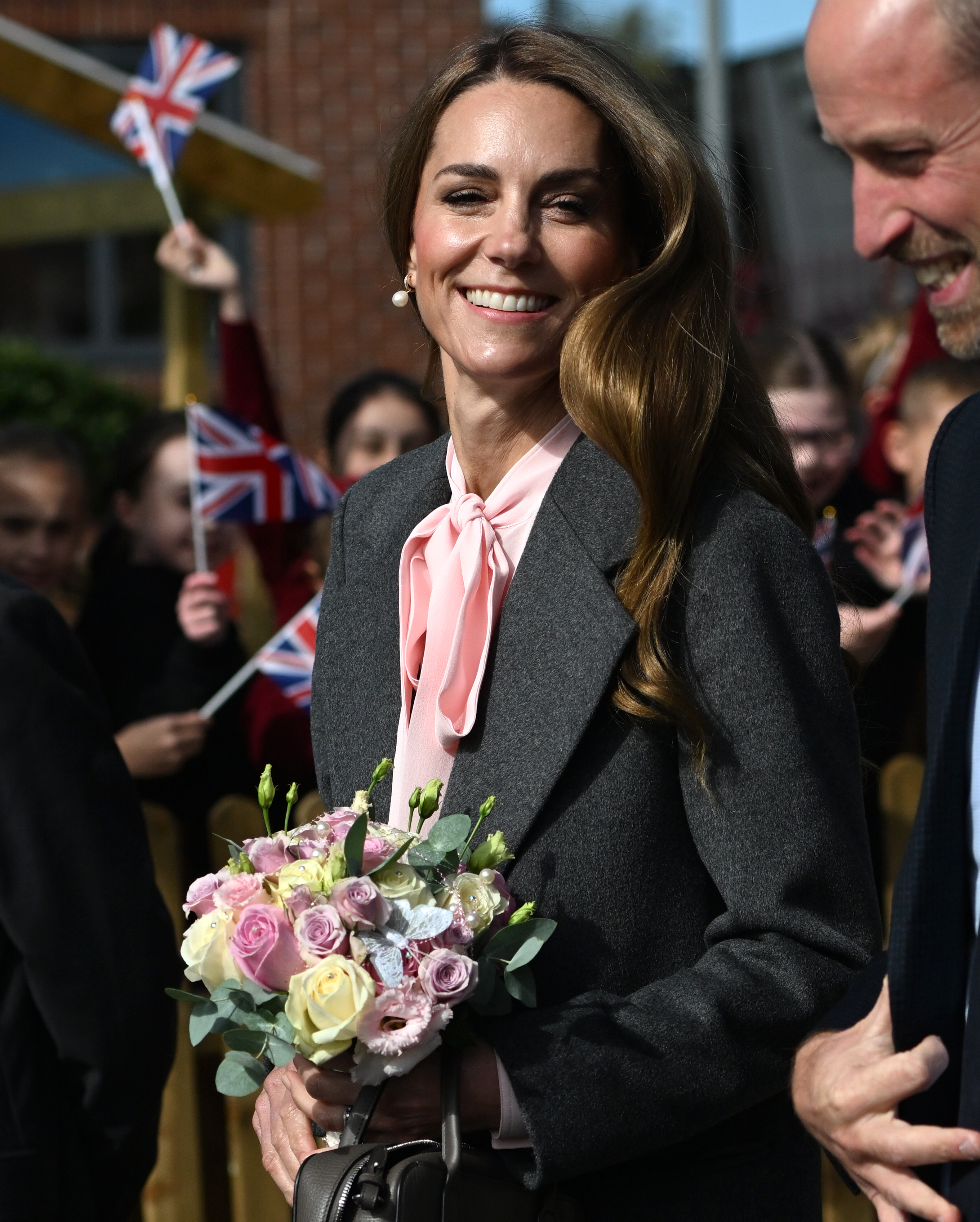 Kate Middleton wearing a gray suit and holding flowers and smiling