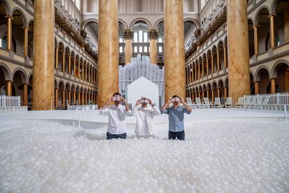 Snarkitecture partners Alex Mustonen, Daniel Arsham, and Ben Porto in their ‘Fun House’ installation at the National Building Museum. The three people are standing in an area filled to waist height with white balls. 