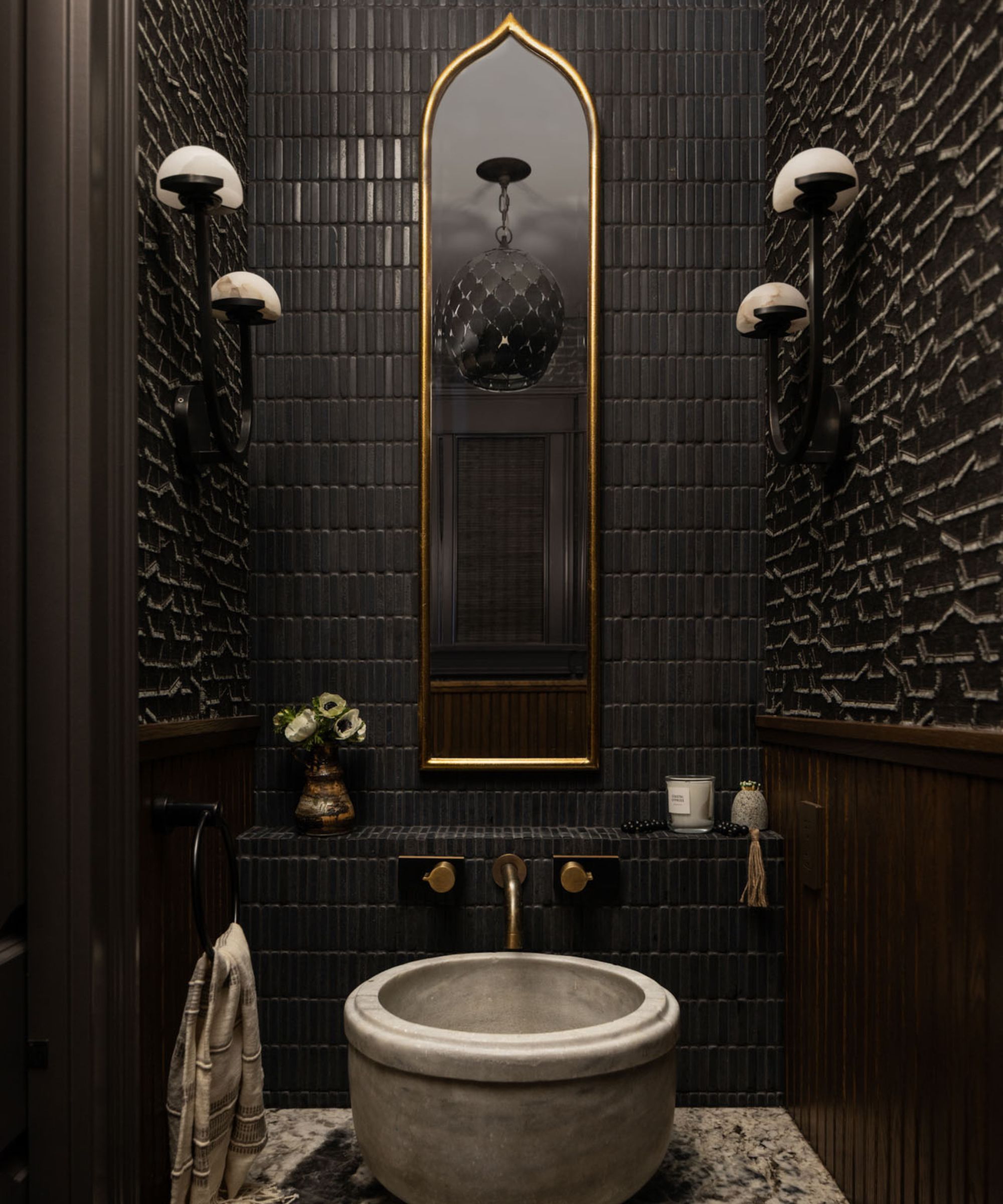 A small powder room decorated with black textured wallpaper, a brass mirror, and a round stone sink