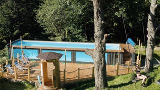 above ground swimming pool made from shipping container with glass side panels, decked area to side and wood and rope fence dividing deck and lawn