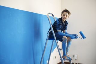 A boy is sitting on a ladder next to a white and blue wall, he has blue paint on his hands, face and legs and is holding a paint roller.