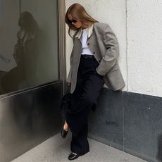 Hanna wears black trousers, white shirt, and aa gray blazer with black shoes and black sunglasses